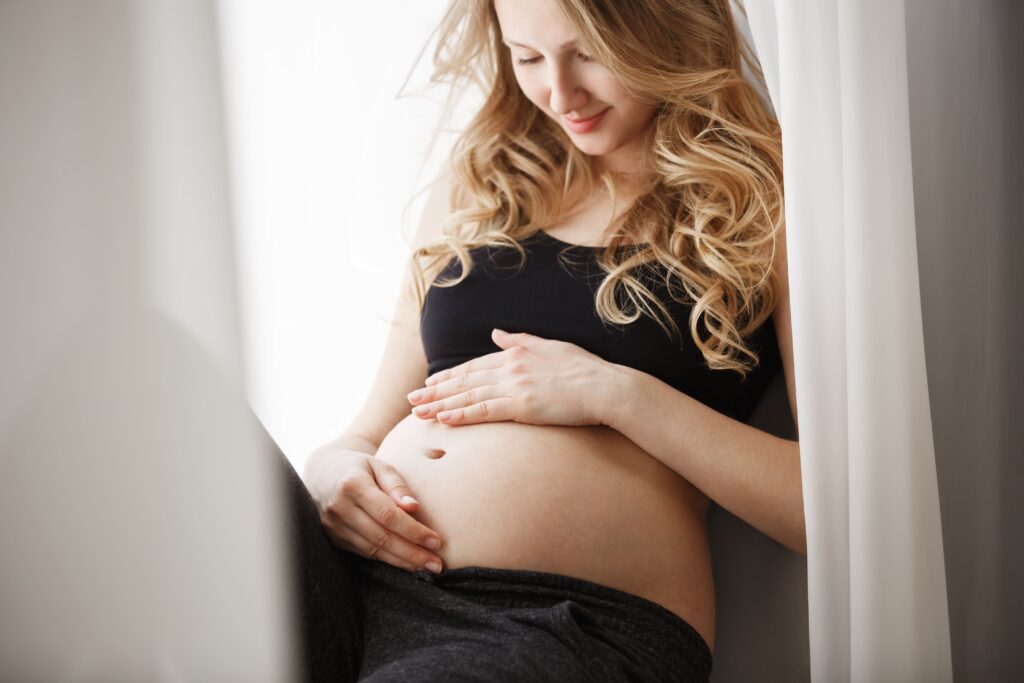 jovem-mãe-grávida-loira-vestida-de-preto-sentado-no-peitoril-da-janela-no-quarto-tocando-e-olhando-para-a-barriga-com-expressão-feliz Mulher grávida sentada na janela, enquanto abraça e toca sua barriga.