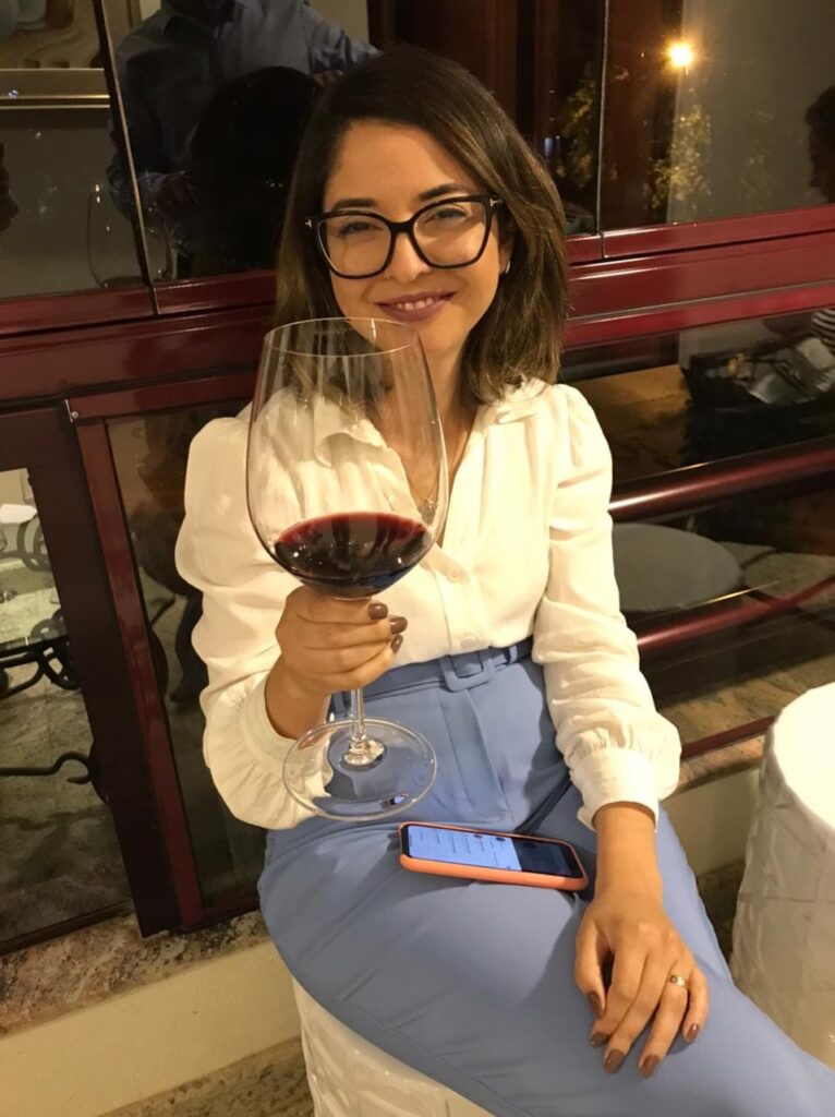 Bianca-sentada-segurando-uma-taça-de-vinho Bianca vestindo uma calça azul clara e blusa branca, enquanto segura uma taça de vinho e sorri para a câmera.