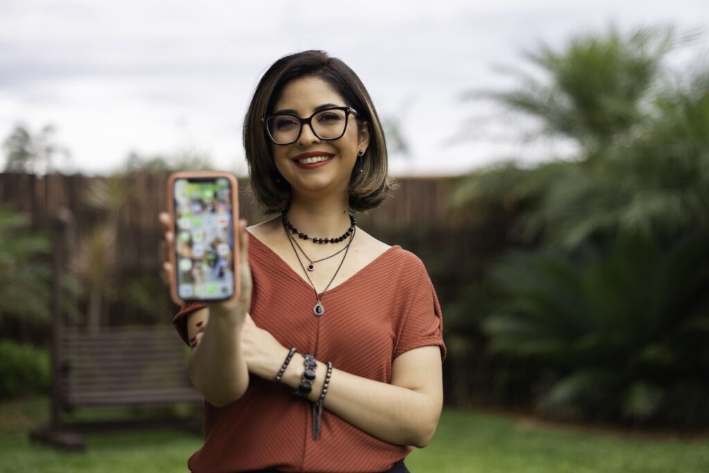 Bianco-sorrindo-segurando-o-celualar-e-mostrando-a-tela-do-celular-para-a-câmera Bianca sorrindo enquanto segura um celular e mostrando a tela para a câmera.